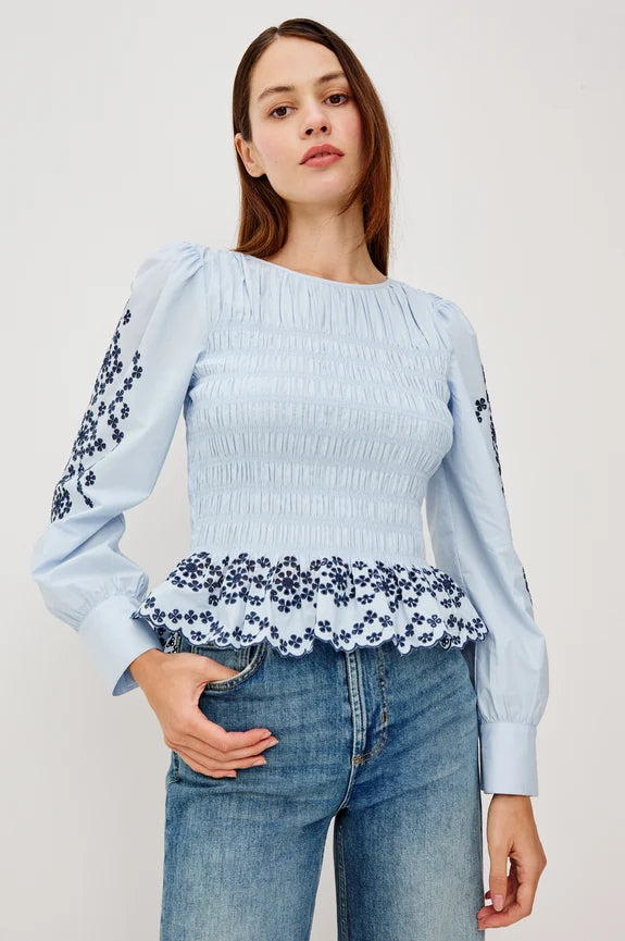 Brunette wearing light blue long-sleeve peplum blouse with navy eyelet embroidery