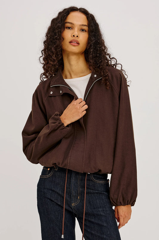 Woman wearing a brown jacket and blue jeans against a plain background